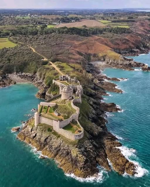 Aerial photo of a ruined stone castle on a rocky cliff juting into turquoise ocean with green fields behind
