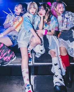 Four young women in colorful punk-style outfits posing together on a stage under blue and pink lights.