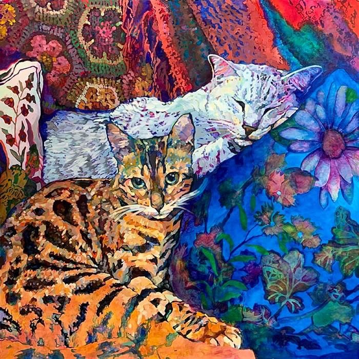 Two cats lying on colorful patterned fabric with floral designs and decorative pillows