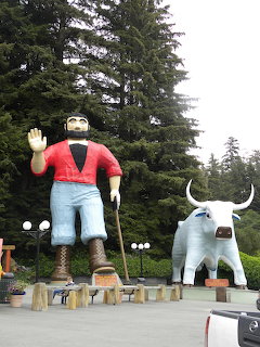Giant Paul Bunyan statue in red shirt and blue jeans next to Babe the Blue Ox statue among evergreen trees