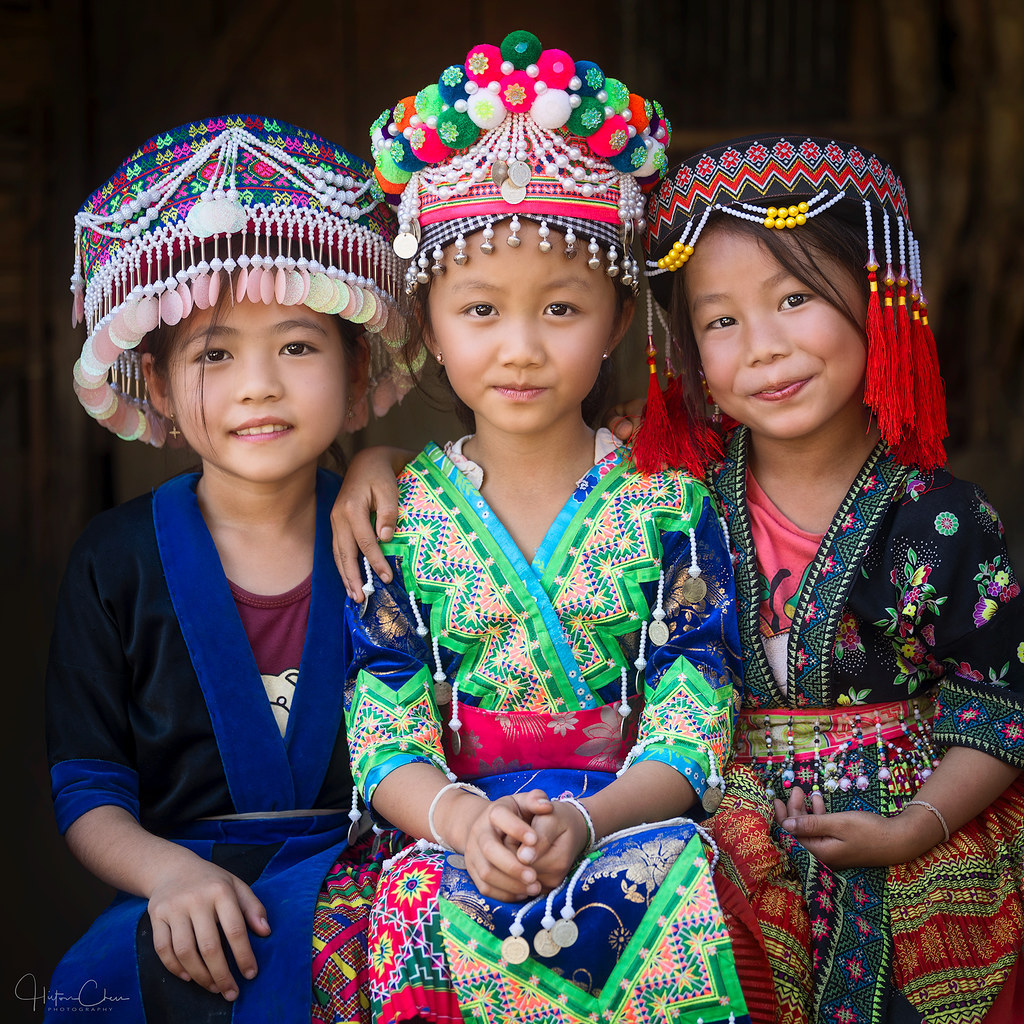 Three smiling girls in vibrant traditional Hmong costumes with decorative silver headdresses