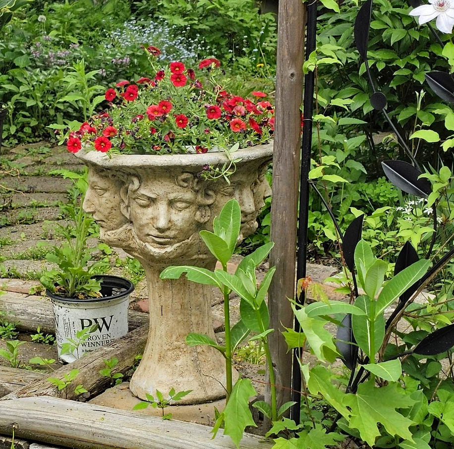 Stone face planter with red flowers in a leafy garden near a brick path