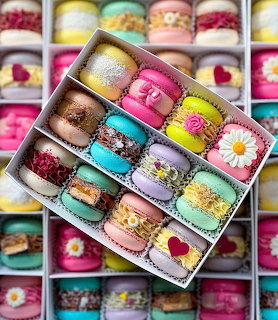 Colorful macarons arranged in rows inside a white box, with more macarons blurred in background