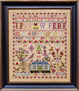 Framed antique needlework sampler with alphabet, house, figures, and floral patterns in red and green on cream linen
