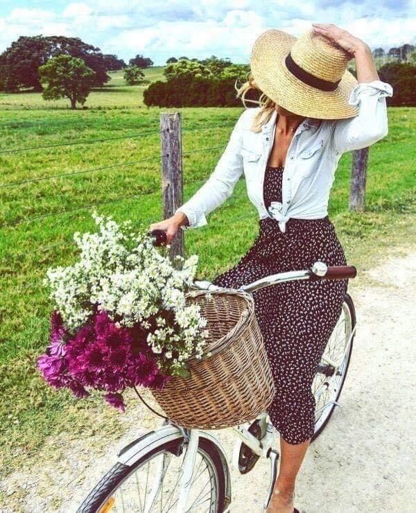 Woman in a straw hat standing with a bicycle and flowers beside a green field