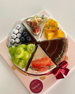 Round cheesecake with varied fruit toppings including blueberries, figs, mango, grapes, and grapefruit in a pink box