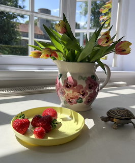 Tulips in a floral jug on a sunny windowsill beside a plate of strawberries