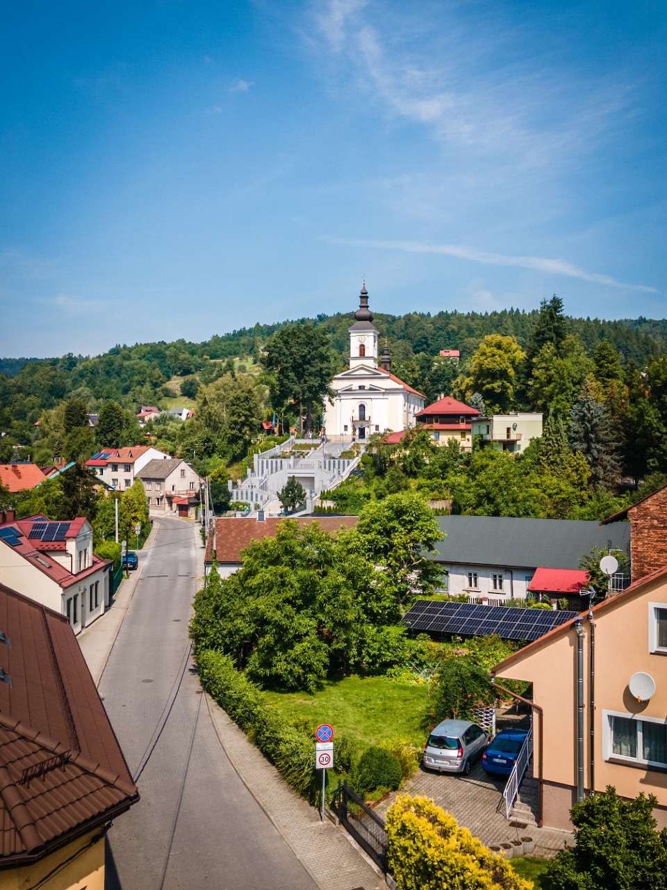 Aerial view of a small village with a white church on a hill, green trees, and residential buildings