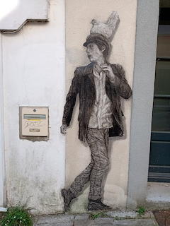 Wall painting of a man walking with a hen on his head, drawn in charcoal style on a white building exterior