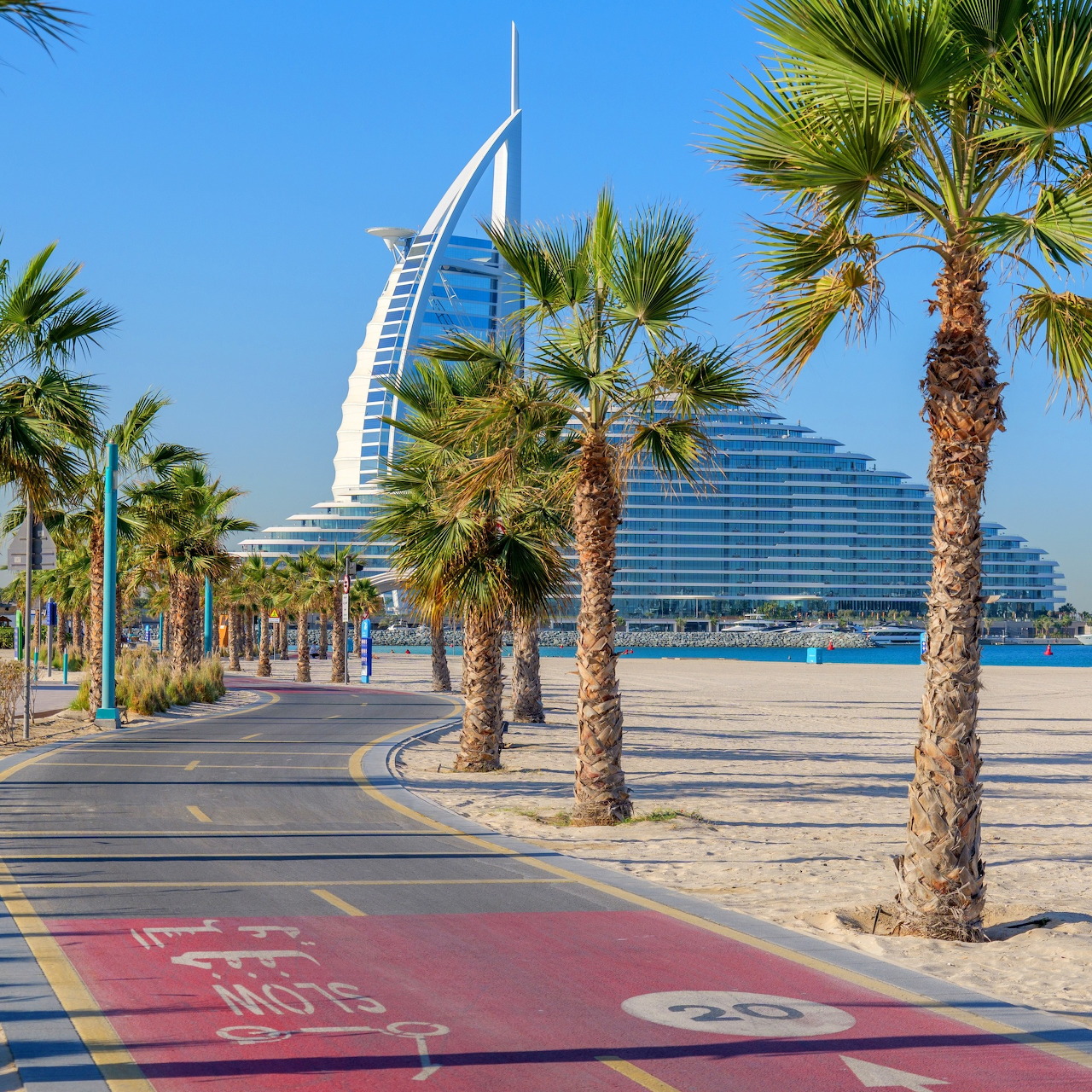 Burj Al Arab hotel with palm trees along Dubai waterfront promenade under clear blue sky