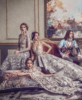 Four women in elaborate silver and grey couture gowns posing in an opulent baroque-style room