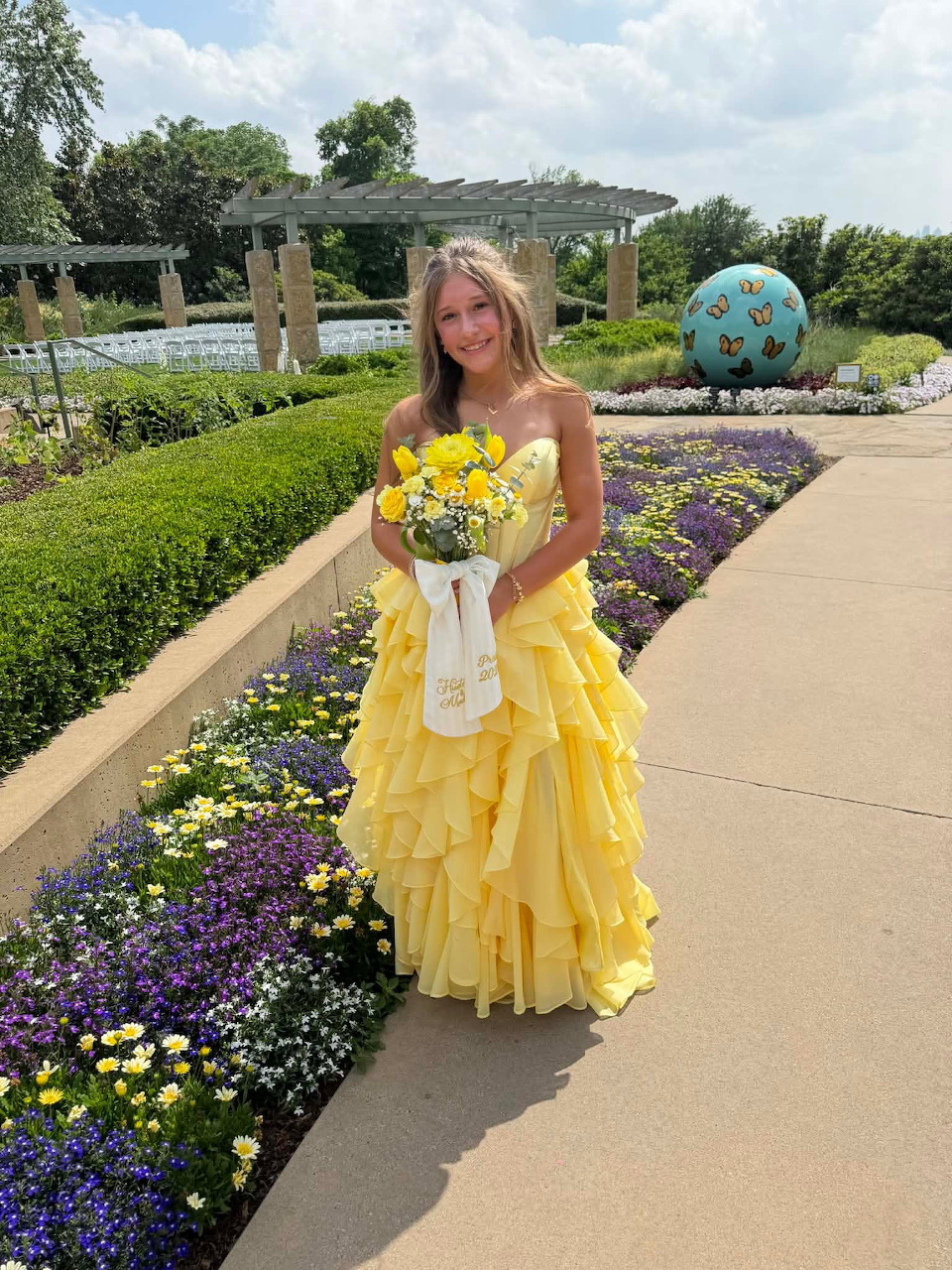 Teenage girl in yellow prom dress holding flowers in a garden with colorful flower beds