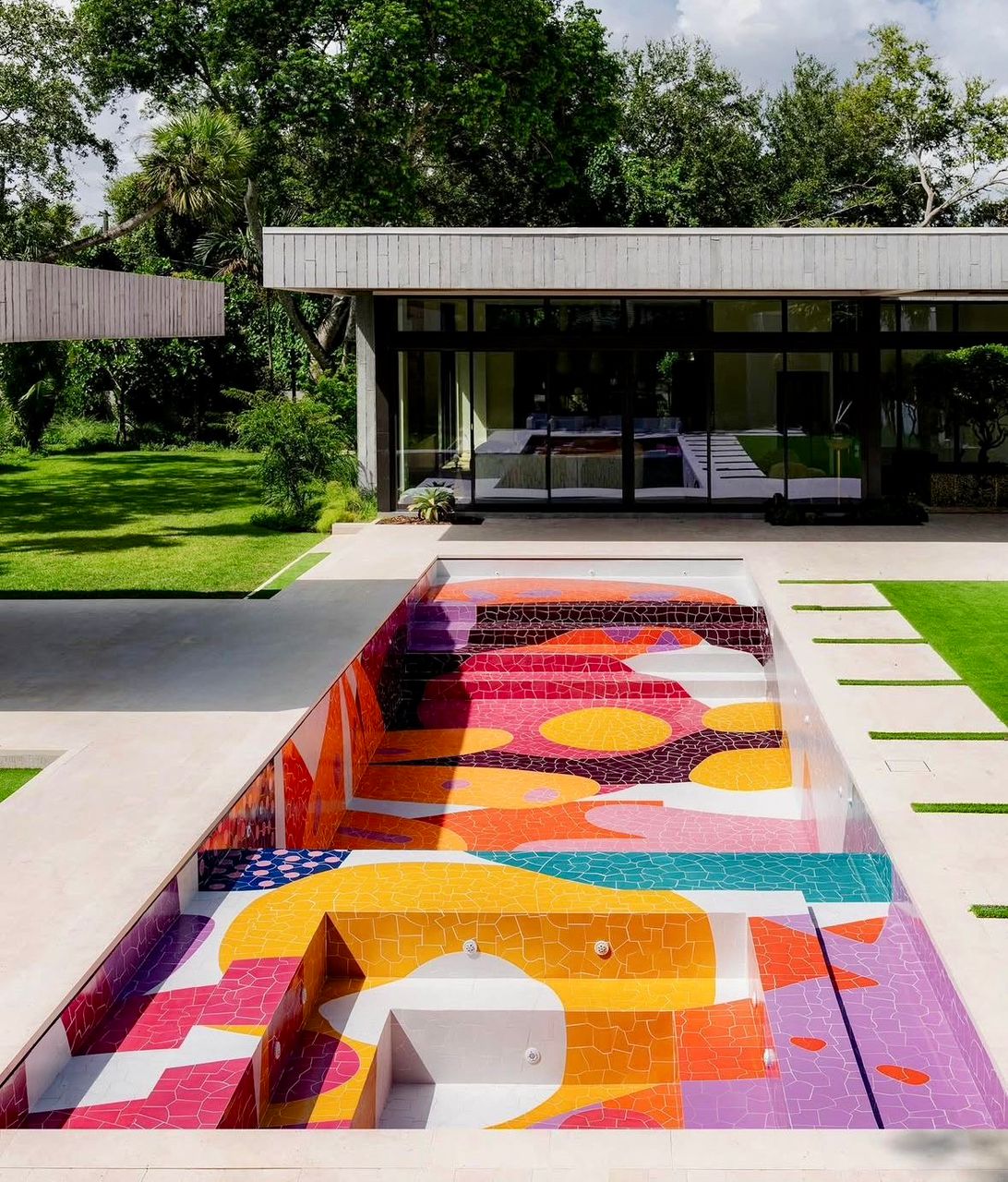 Modern swimming pool with colorful abstract mosaic tile bottom surrounded by green lawn and contemporary architecture
