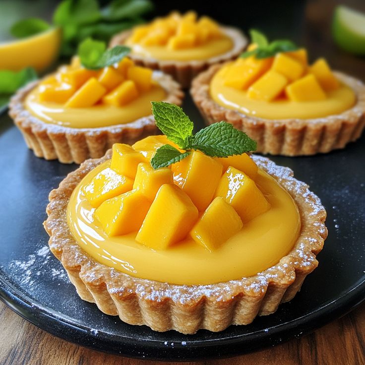 Three golden mango tarts with fluted crusts, yellow custard filling, fresh mango pieces, and mint garnish on black slate