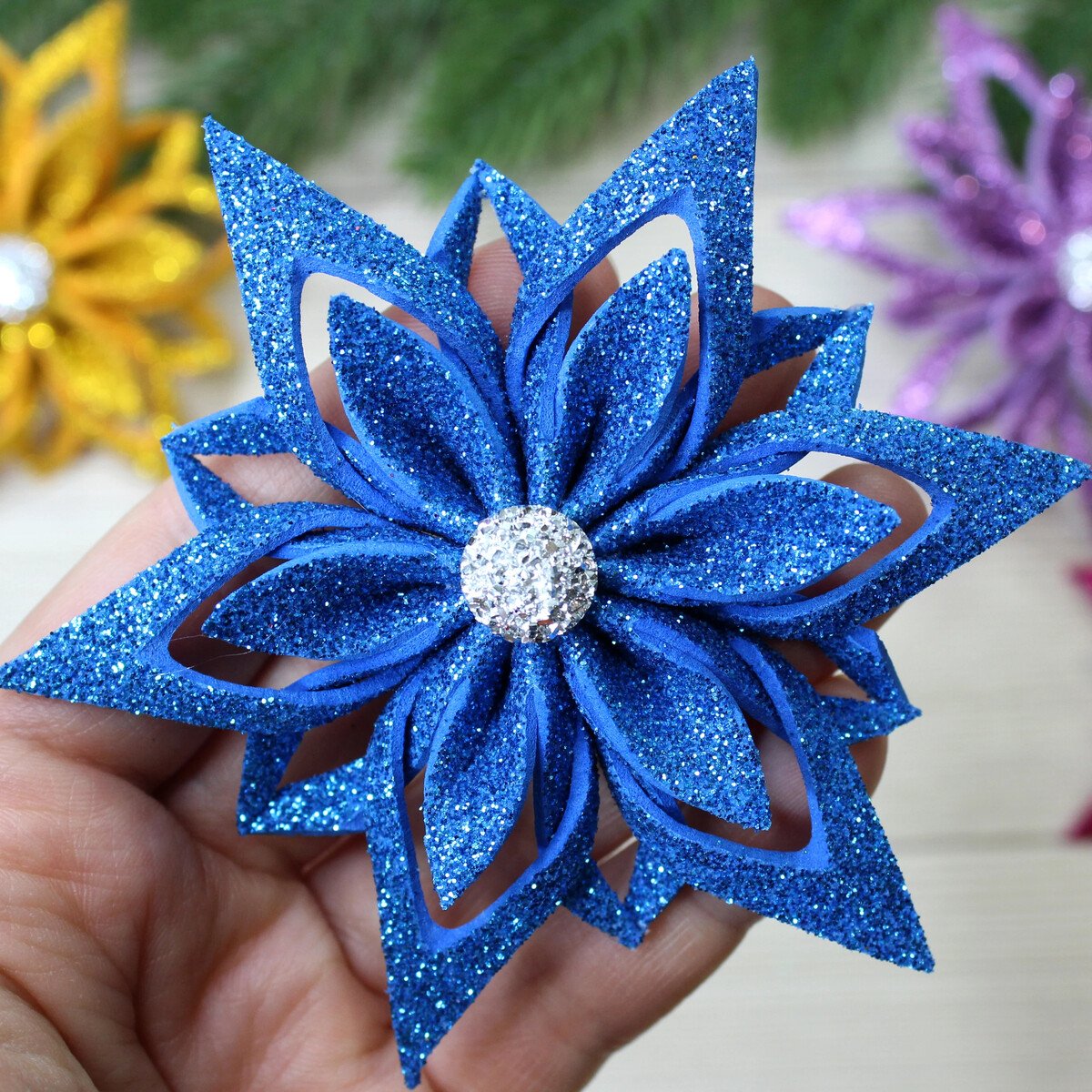 Hand holding a blue glitter snowflake ornament with layered petal shapes