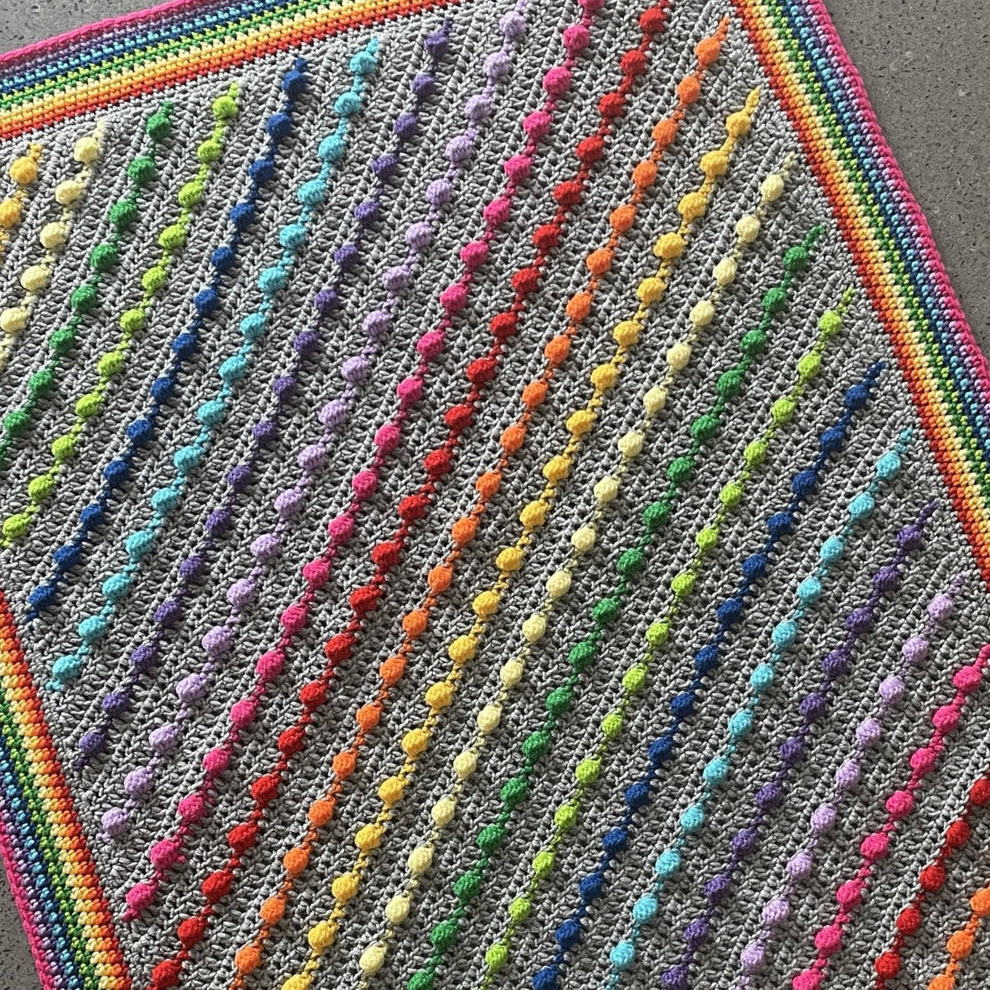 Colorful crocheted blanket with diagonal bobble stitch stripes in rainbow colors on gray background with rainbow border