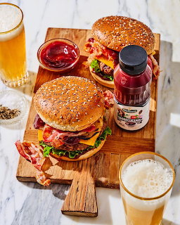 Two bacon cheeseburgers on sesame buns with condiments and beer glasses on wooden board