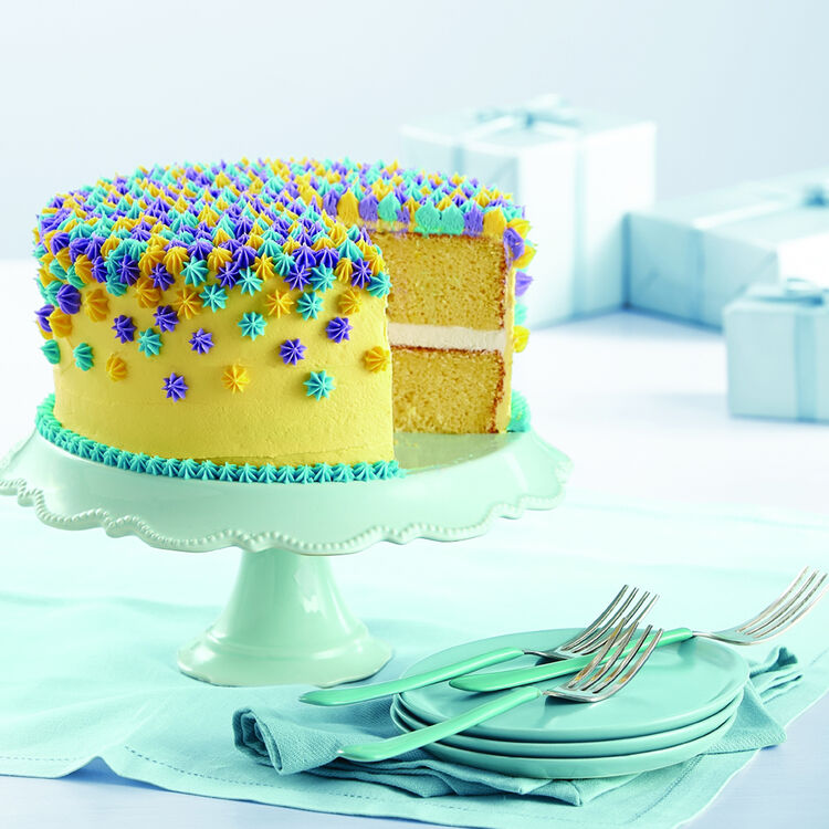 Yellow layer cake with white frosting and multicolored star sprinkles on mint green cake stand with plates and forks