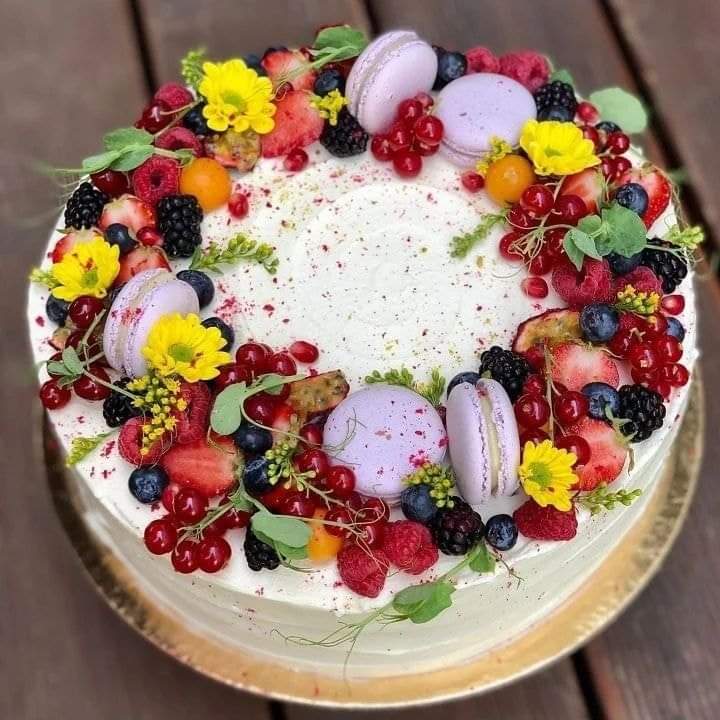 Round white cake topped with berries, macarons, and edible flowers in a decorative wreath pattern
