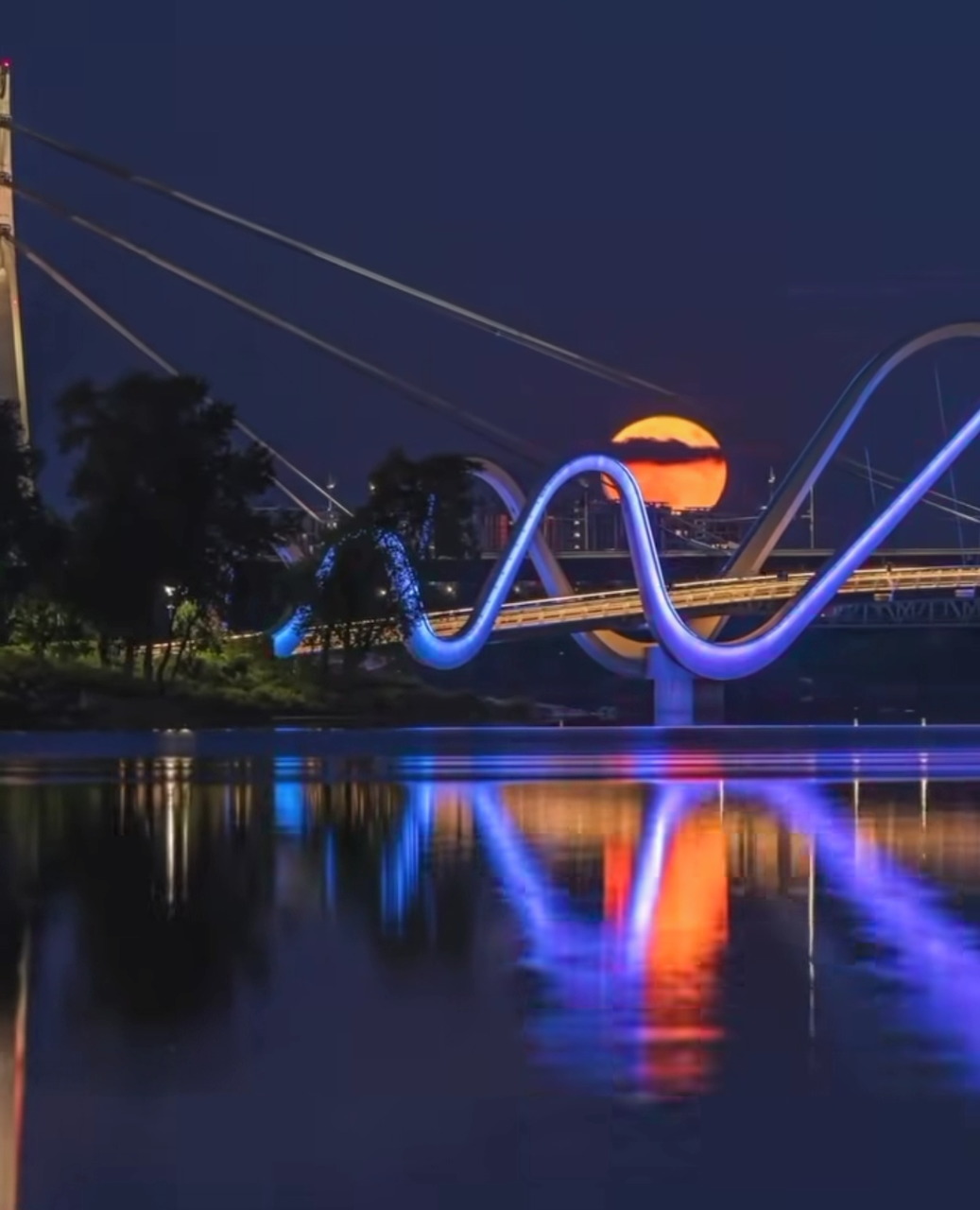Glowing wavy bridge reflected in calm water at night with a large orange moon behind it