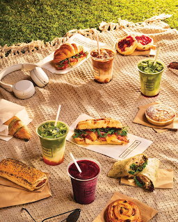 Assorted sandwiches, pastries, and colorful drinks spread on a woven picnic blanket outdoors in sunlight