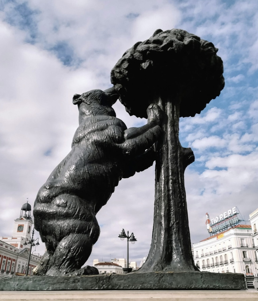 Bronze statue of bear reaching up to tree, Madrid's coat of arms symbol, under cloudy sky