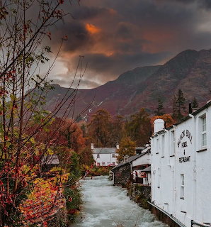 White village buildings along a stream with autumn trees and dark mountains under dramatic orange clouds