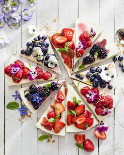 Cheesecake slices topped with assorted fresh berries and edible flowers arranged in a circular pattern on white wood