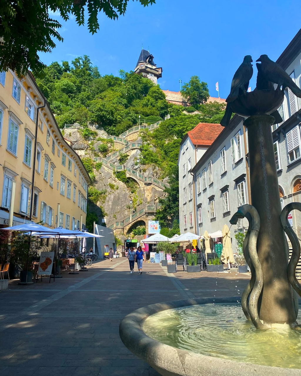 Graz clock tower on hilltop above European town square with fountain and cafes