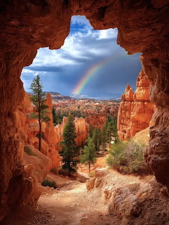 Rainbow arching over red rock canyon viewed through natural stone arch with pine trees and hoodoos