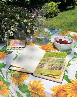 Open watercolor sketchbook on sunflower tablecloth in garden with paint set, flowers, and berry bowl