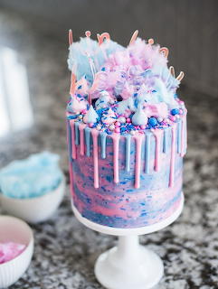 Pastel drip cake topped with cotton candy, macarons, and sprinkles on a cake stand