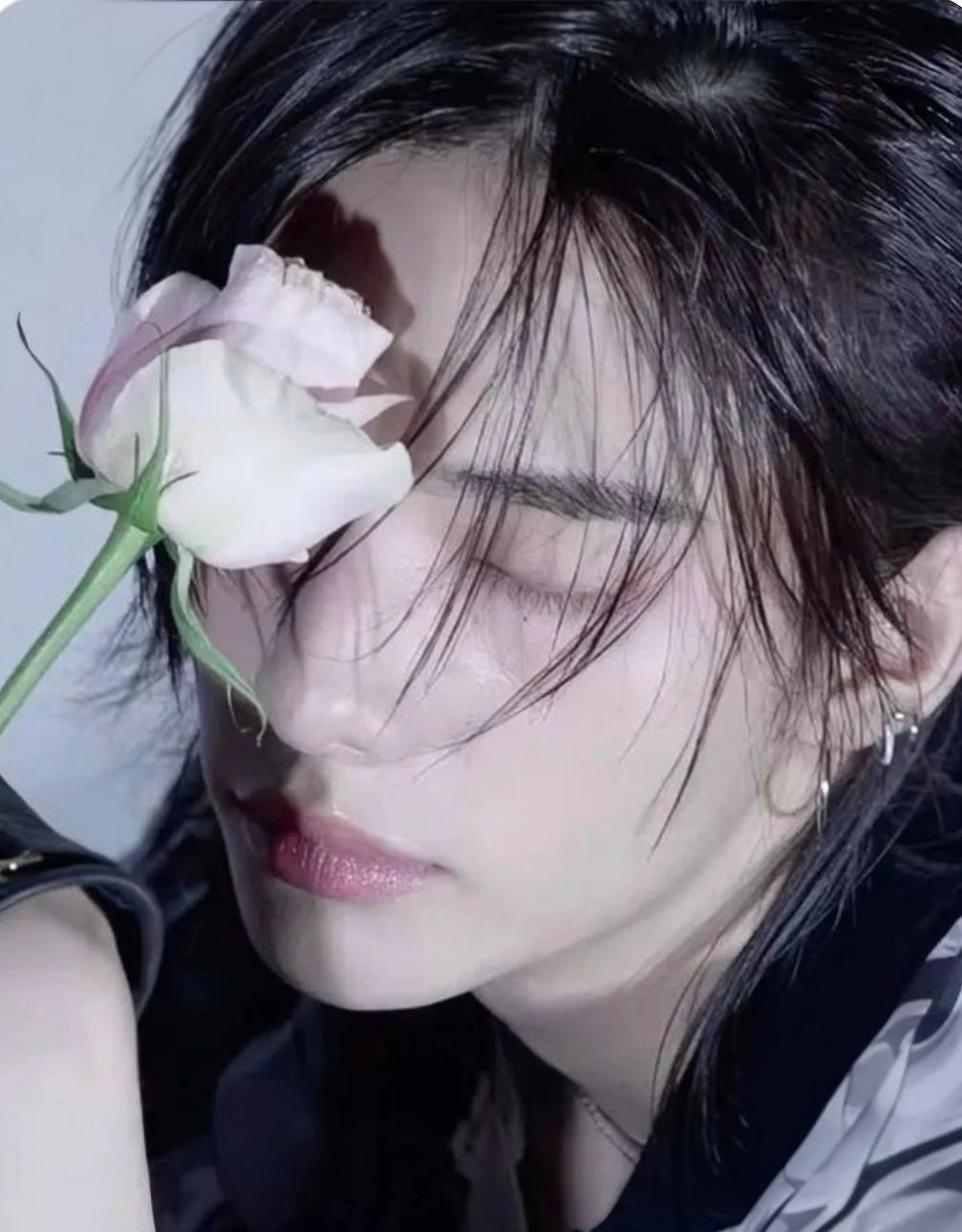 Close-up portrait of a person with dark hair and a pale pink rose covering one eye