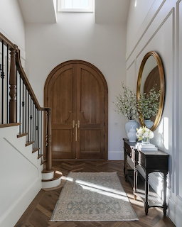 Bright foyer with arched wooden door, staircase, console table, round mirror, and rug