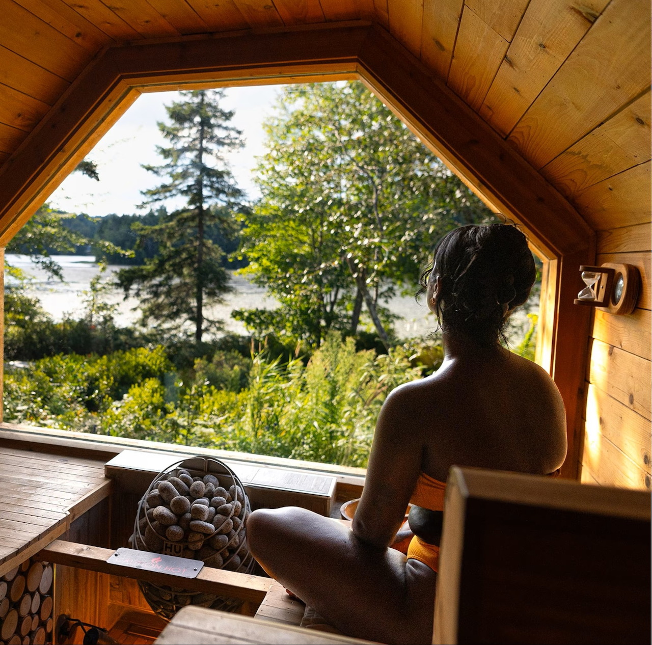 Interior of wooden sauna with arched window showing lake and forest view, sauna stove visible in foreground