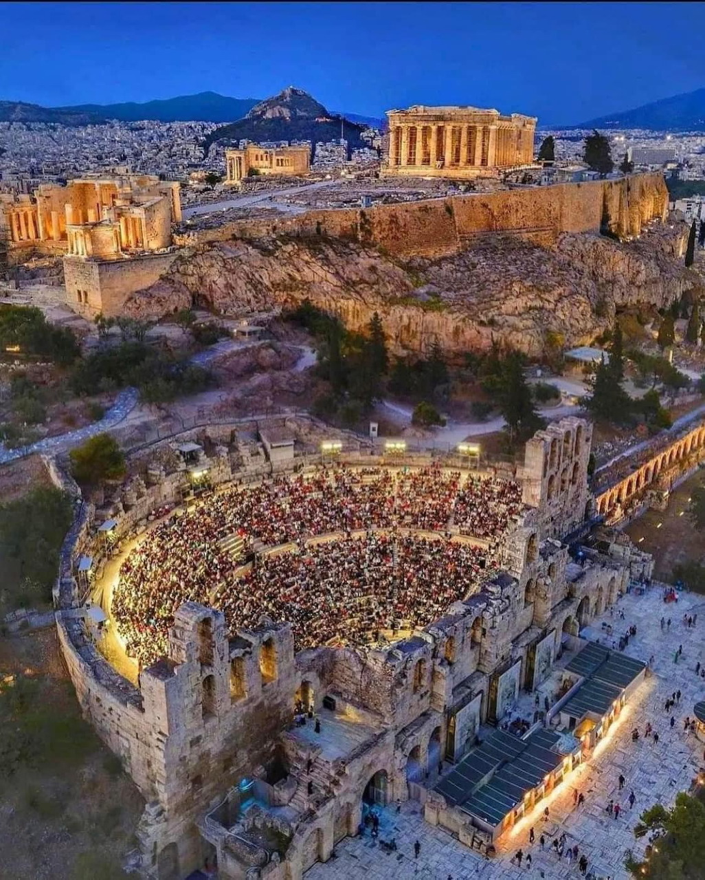 Aerial view of illuminated ancient Herodion theater at Acropolis with Parthenon and Athens cityscape at dusk