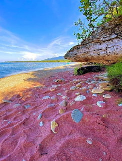 Red garnet sand beach with scattered pebbles, rocky cliff, blue water, and bright sky