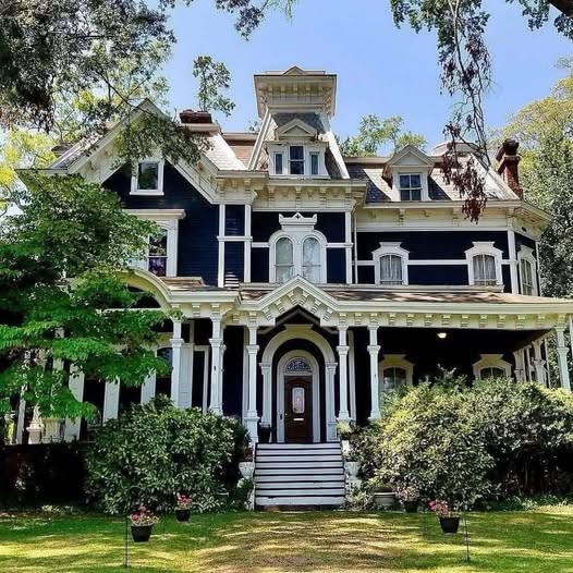 Victorian mansion with white ornamental trim, central tower, and wraparound porch surrounded by greenery
