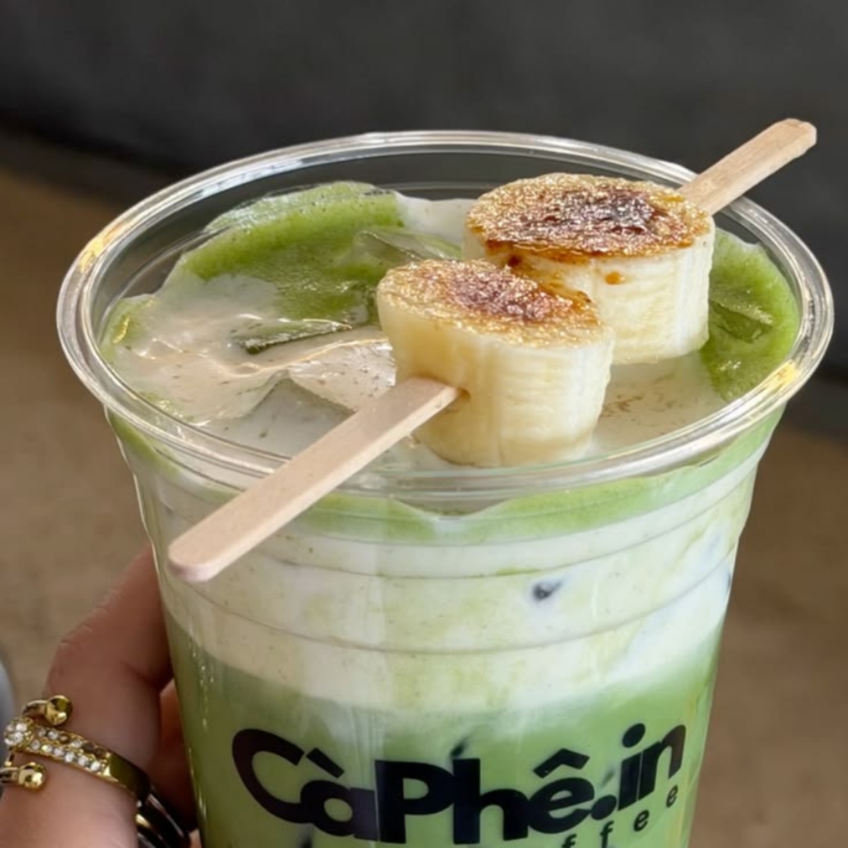 Iced matcha drink in a clear cup with two banana slices on wooden sticks on top