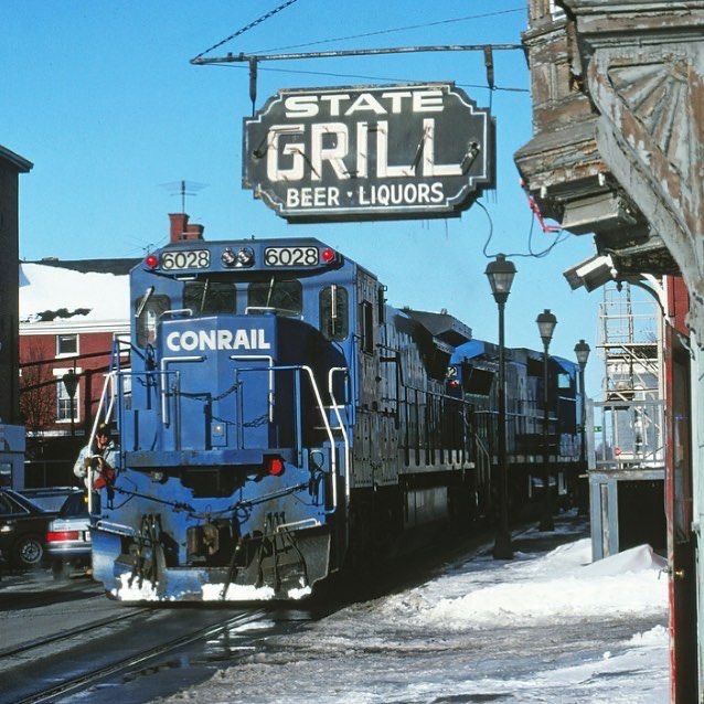 Blue Conrail train moving through a snowy town street under a State Grill sign