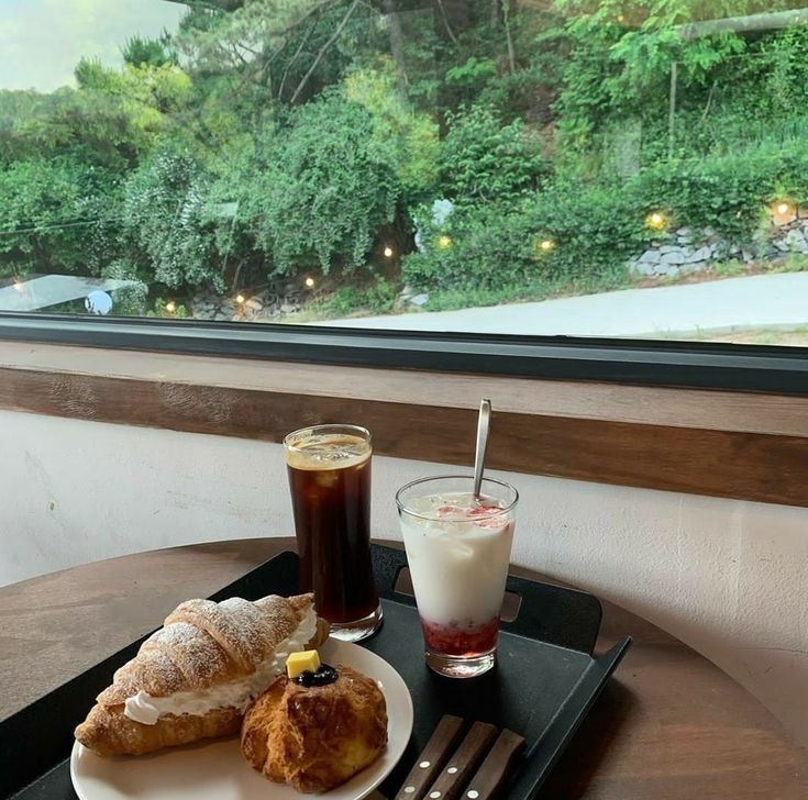 Tray with coffee drinks, croissant, and pastry on a table by a window overlooking greenery