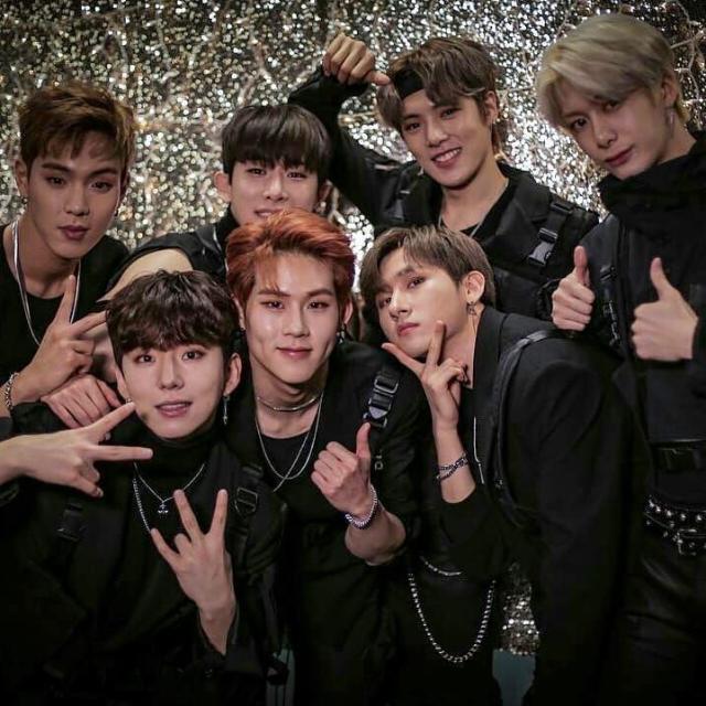 Seven young men in black clothing posing together making peace signs and other hand gestures with glittery background