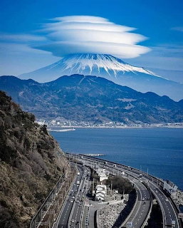 Mount Fuji with a lenticular cloud above coastal hills and a curving seaside highway