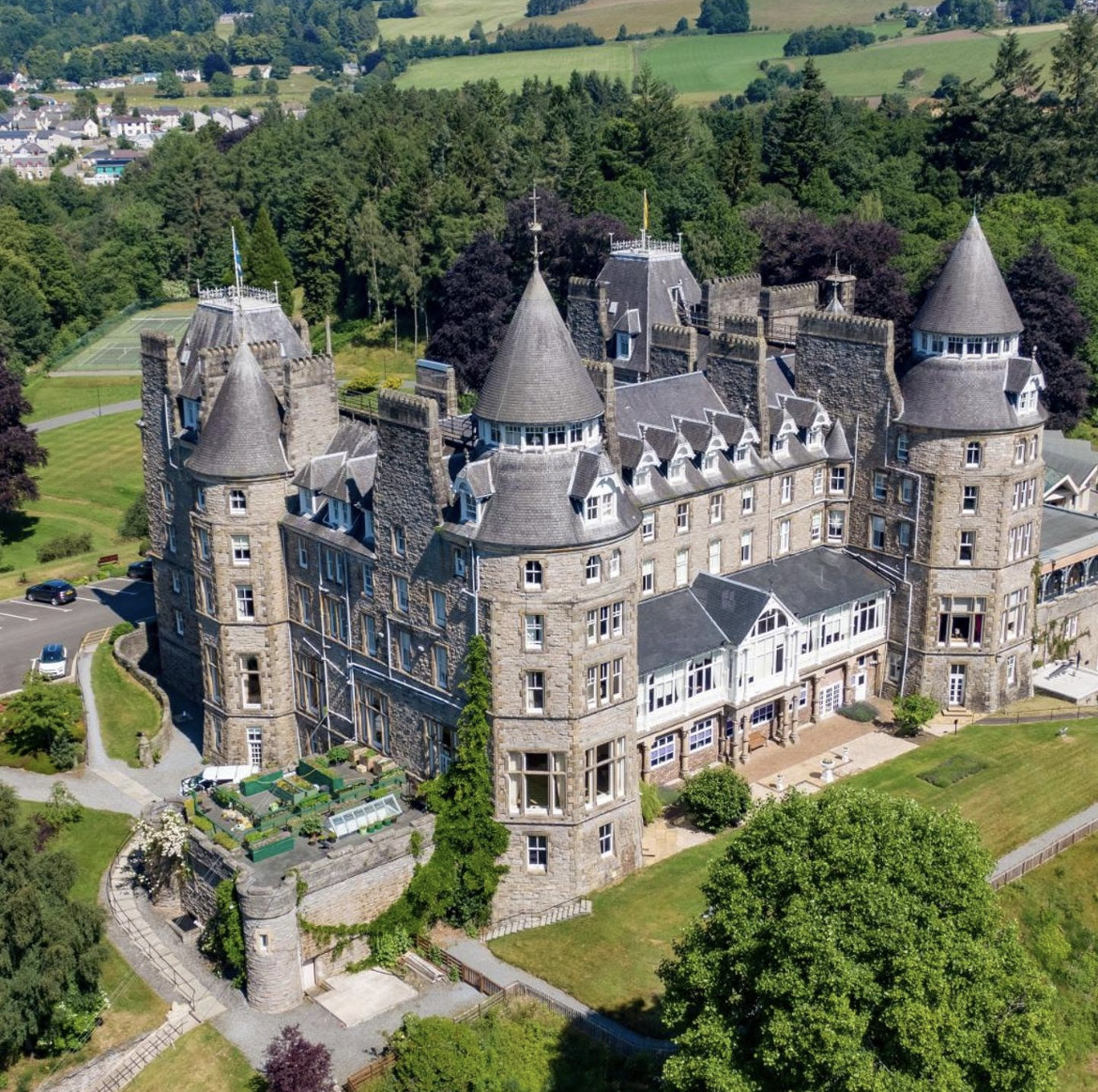 Aerial view of a large stone castle with round towers and pointed roofs surrounded by green fields and forests