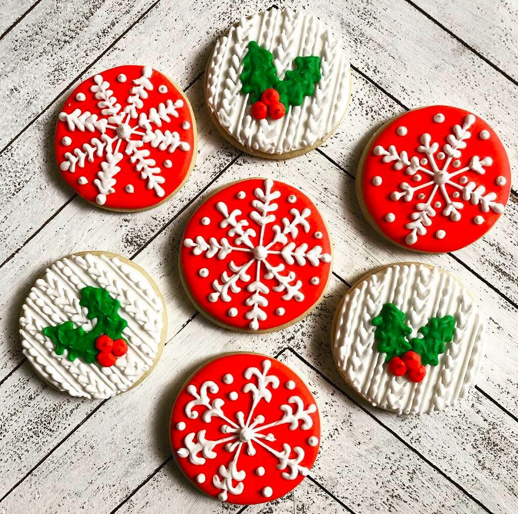 Six decorated Christmas cookies with red snowflake icing and white holly designs on a wooden background.