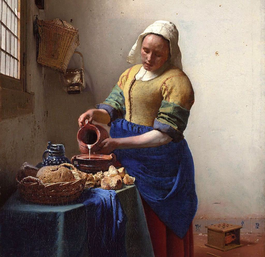 Woman in blue and yellow dress pouring milk from ceramic jug in rustic kitchen