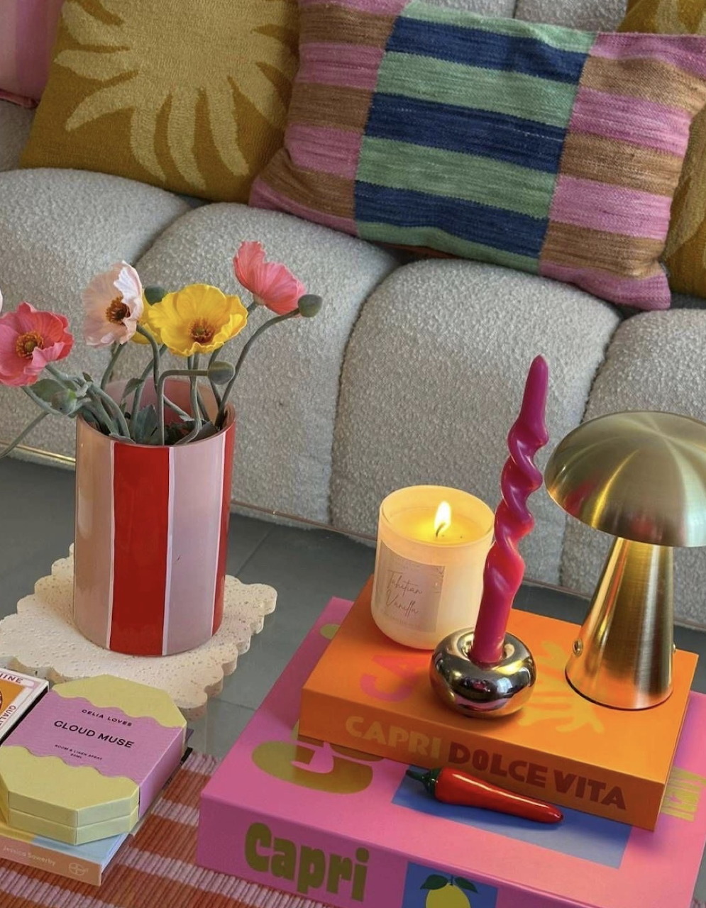 Coffee table with striped flower vase, lit candle, pink taper candle, gold lamp, and colorful books on a pink surface