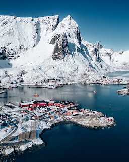 Aerial view of a snowy Arctic fishing village with red buildings beneath dramatic snow-covered mountains
