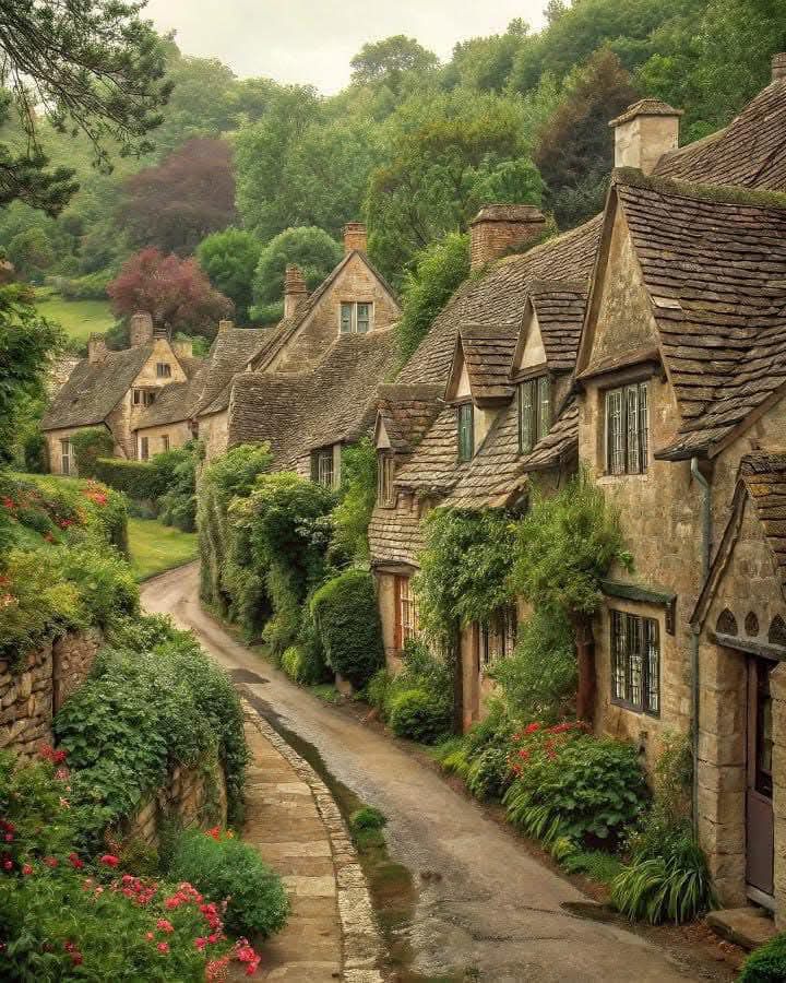 Stone cottages along a narrow village lane in Bibury, Gloucestershire, with greenery and flowers