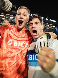 Two soccer players celebrating intensely at night, one in orange goalkeeper kit and one in white jersey